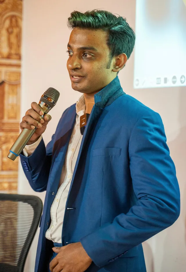 Vairavan presenting with a handheld microphone at the Kamala Club